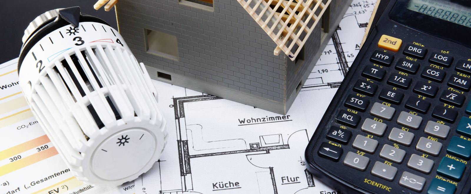 Symbolbild für Altbausanierung mit einem Modell eines Hauses, einem Heizungsthermostat und einem Taschenrechner, die auf einem Grundriss liegen