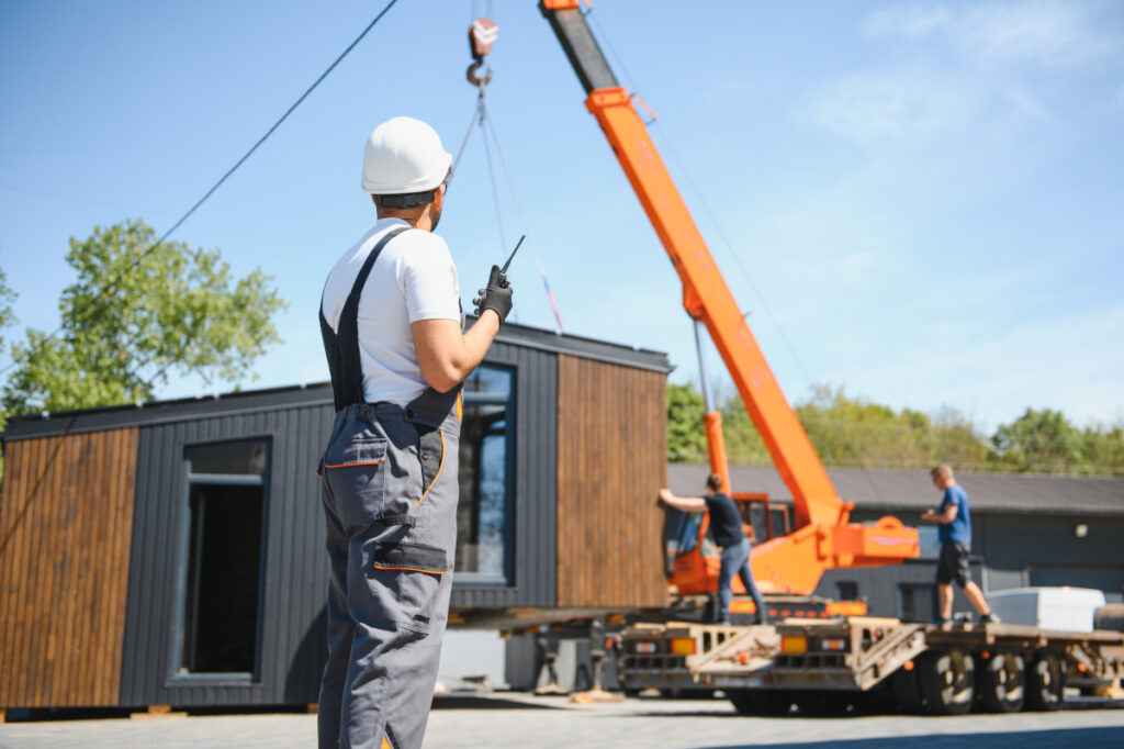 Bauarbeiter mit Funkgerät vor einem Kran, der ein Modulteil eines Modulhauses auf einen LKW lädt