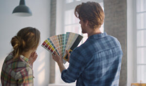 Ein junges Paar steht in einem Raum und halt eine Farbpalette für Wandfarben hoch. Symbolbild für kreative Raumaufteilung sowie Decken- und Wandgestaltung. 