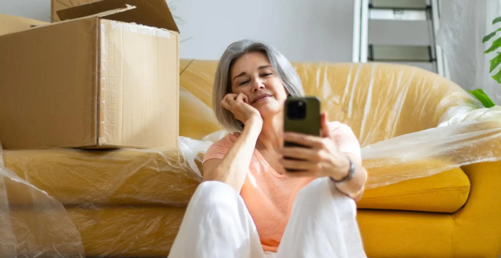 Ältere Frau mit Smartphone hockt vor Couch und Umzugskartons